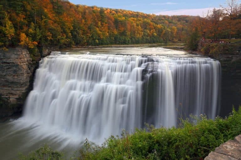 A river, surrounded by fall foliage, drops into a high multi-level waterfall in Letchworth Park in the Finger Lakes region